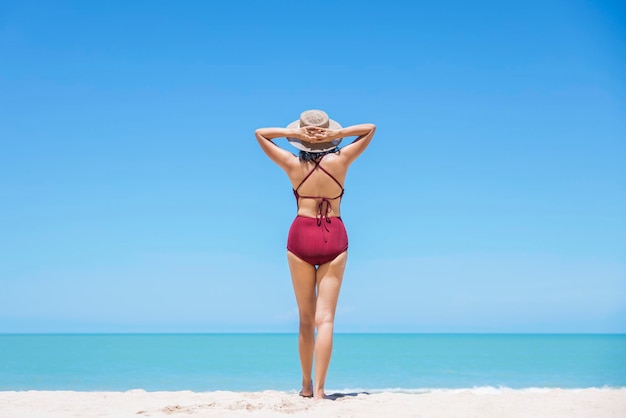 Młode kobiety w bikini i słomkowym kapeluszu stoją na tropikalnej plaży, ciesząc się widokiem oceanu na plaży