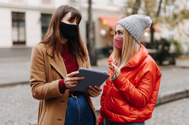 Młode kobiety w maskach medycznych, stojąc na ulicy miasta i trzymając cyfrowy tablet. Koleżanki spędzają czas na świeżym powietrzu z nowoczesnym urządzeniem.