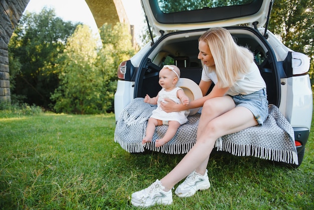 Młode małżeństwo i córka odpoczywają na łonie natury. Kobieta i dziewczyna siedzą na otwartym bagażniku samochodu.