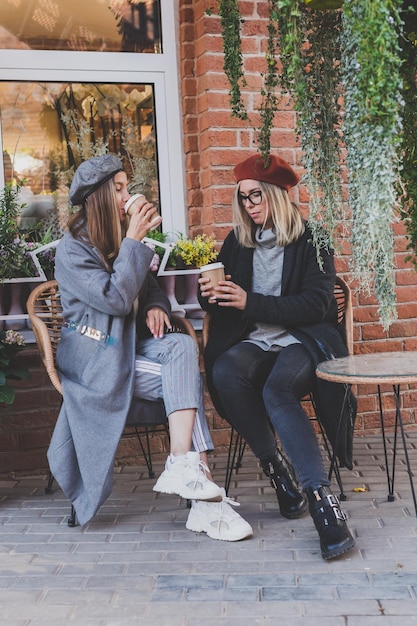 Młode najlepsze przyjaciółki rozmawiają ze sobą, omawiając ciekawe tematy w czasie wolnym hipster dziewczyny żartują i bawią się razem, ciesząc się rekreacją przy kawie na miejskim tle