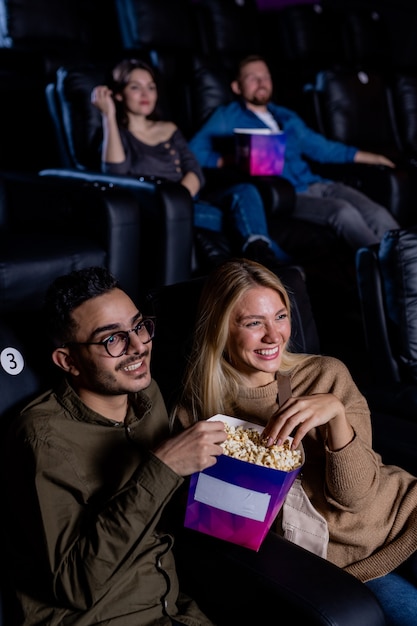 Młode randki z pudełkiem popcornu siedzą w ciemnym kinie i patrzą na duży ekran podczas oglądania filmu w wolnym czasie