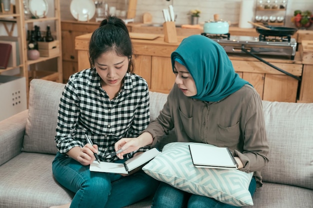 Młode studentki przygotowujące się do egzaminu na kanapie. wieloetniczne współlokatorki dziewczyny siedzące na kanapie, omawiając projekt i pisząc na księdze notatek. piękna muzułmańska dama w hidżabie uczy przyjaciela zadanie domowe