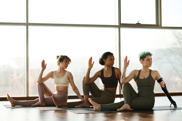 Młode zdrowe kobiety siedzące z rzędu na macie do ćwiczeń i wykonujące ćwiczenia jogi