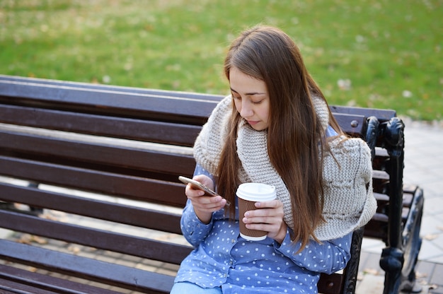 Młodej dziewczyny obsiadanie na ławce w jesień parku pije kawę i opowiada na smartphone