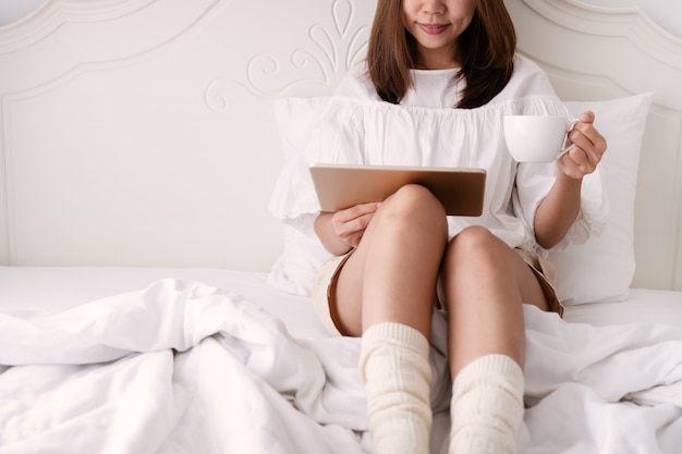 Młodej pięknej brunetki włosiana kobieta używa pastylkę i pijący kawę podczas gdy siedzący na łóżku w ranku. bliska, Leniwy dzień wolny koncepcja