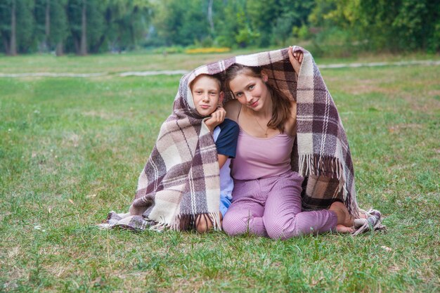 Młodsza Siostra I Brat Z Piegami Na Twarzach Stoją W Parku Okryci Kratą, Rozgrzewają Się, Uśmiechają I Patrzą W Kamerę.