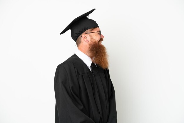 Młody absolwent uniwersytetu czerwonawy mężczyzna na białym tle śmiejący się w pozycji bocznej