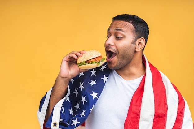 Młody african american indian czarny człowiek jedzenie hamburgerów na białym tle nad żółtym tle z amerykańską flagą. Pojęcie diety.