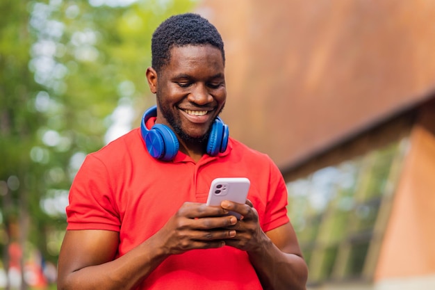 Młody afro amerykański mężczyzna ze słuchawkami na szyi za pomocą smartfona w letnim parku