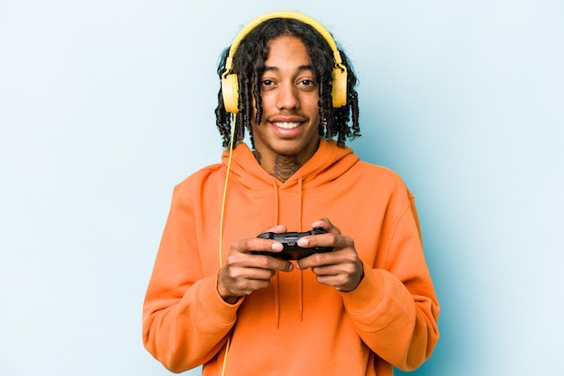 Młody Afroamerykanin bawiący się kontrolerem gier wideo na niebieskim tle szczęśliwy uśmiechnięty i wesoły