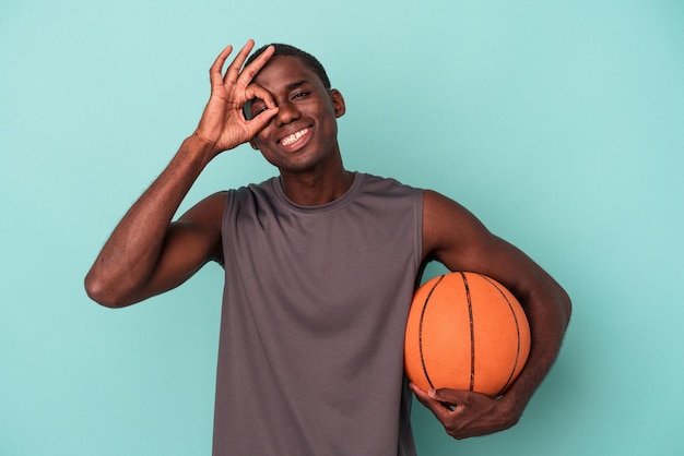 Młody Afroamerykanin gra w koszykówkę na białym tle na niebieskim tle podekscytowany, trzymając ok gest na oko.