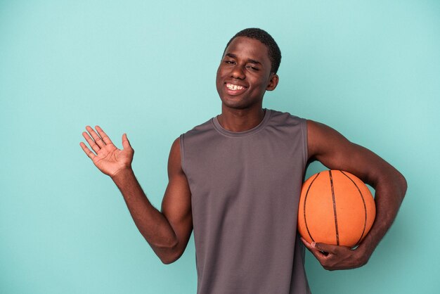 Młody Afroamerykanin gra w koszykówkę na białym tle na niebieskim tle, pokazując miejsce kopii na dłoni i trzymając inną rękę w pasie.