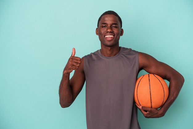 Młody Afroamerykanin gra w koszykówkę na białym tle na niebieskim tle, uśmiechając się i podnosząc kciuk w górę