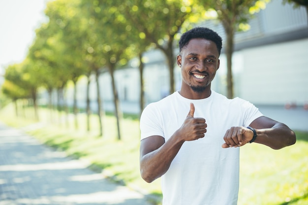 Młody Afroamerykanin, podczas porannego joggingu w białej koszulce w parku, zadowolony z wyniku treningu zaangażowanego w aktywny tryb życia