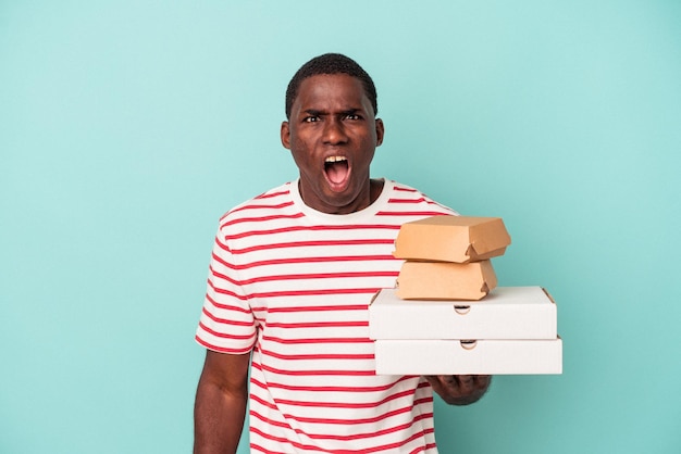 Młody Afroamerykanin posiadający pizze i hamburgery na białym tle na niebieskim tle krzyczy bardzo zły i agresywny.