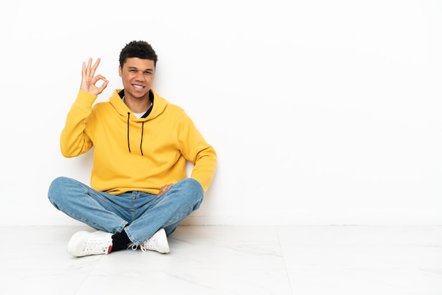 Młody Afroamerykanin siedzący na podłodze na białym tle pokazujący znak ok palcami