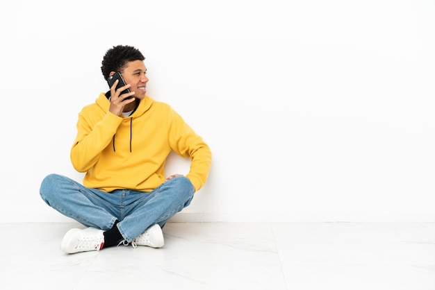 Młody Afroamerykanin siedzi na podłodze na białym tle, prowadząc rozmowę z telefonem komórkowym z kimś