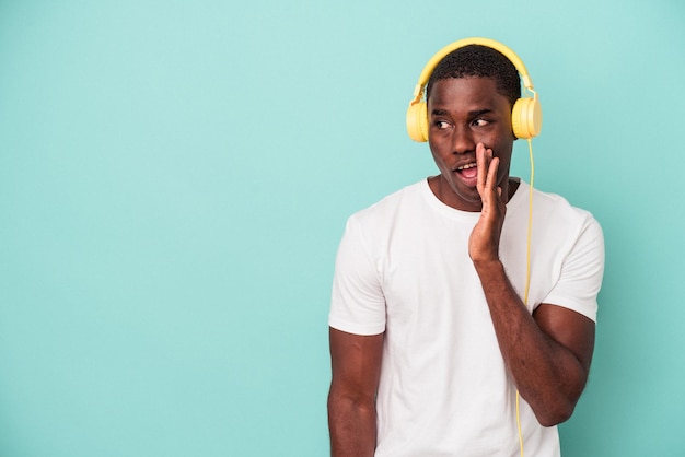 Młody Afroamerykanin słuchający muzyki odizolowanej na niebieskim tle mówi tajne gorące wiadomości o hamowaniu i odwraca wzrok
