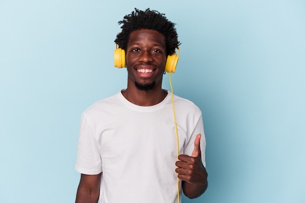 Młody Afroamerykanin słuchający muzyki odizolowanej na niebieskim tle, uśmiechający się i podnoszący kciuk w górę