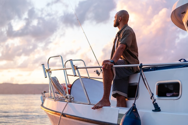 Młody afroamerykanin stojący z wędką na żaglówce łowiący ryby na otwartym morzu o zachodzie słońca