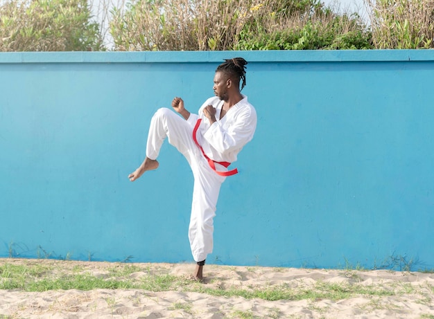 Młody Afroamerykanin trenuje taekwondo na świeżym powietrzu, kopiąc na niebieskim tle