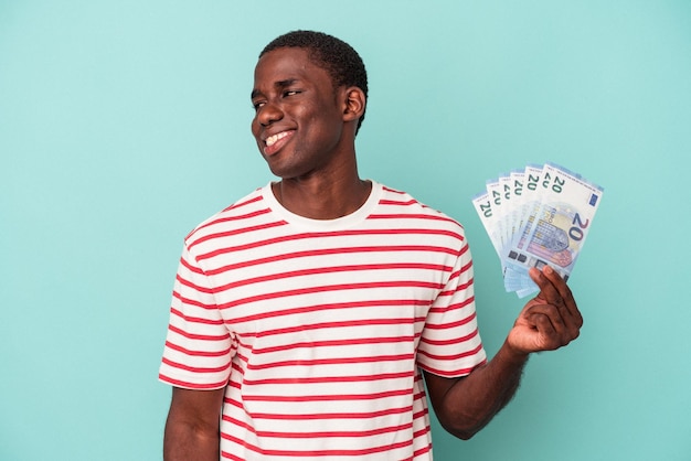 Młody Afroamerykanin trzymający banknoty na białym tle na niebieskim tle wygląda na uśmiechniętego, wesołego i przyjemnego.