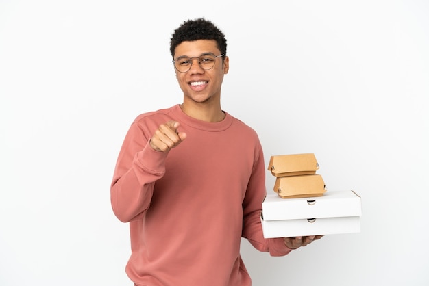Młody Afroamerykanin trzymający burgera i pizze na białym tle zaskoczony i wskazujący przód