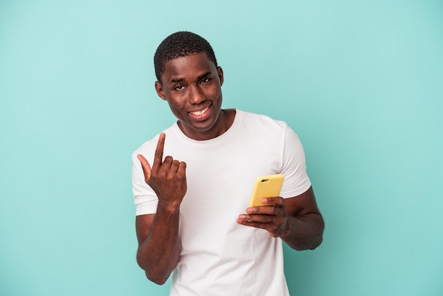 Młody Afroamerykanin trzymający telefon komórkowy na białym tle na niebieskim tle, wskazując palcem na ciebie, jakby zapraszając do siebie.