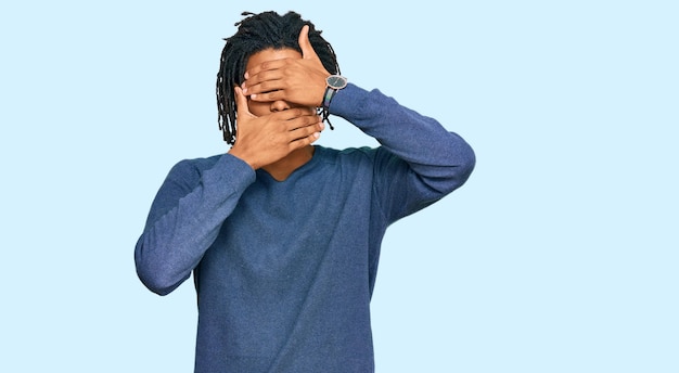 Młody afroamerykanin ubrany w swobodny zimowy sweter zakrywający oczy i usta rękami zaskoczony i zszokowany ukrywaniem emocji