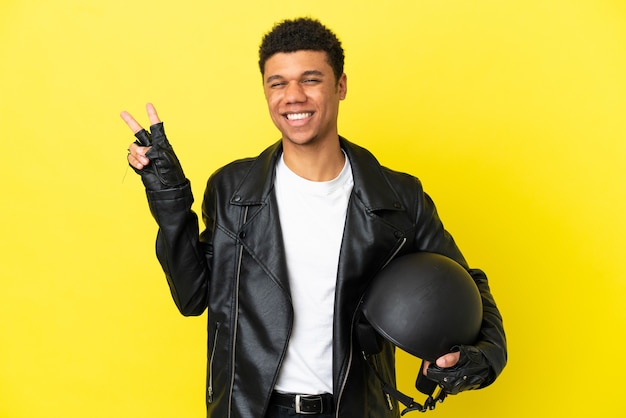 Młody Afroamerykanin w kasku motocyklowym na żółtym tle uśmiechający się i pokazujący znak zwycięstwa