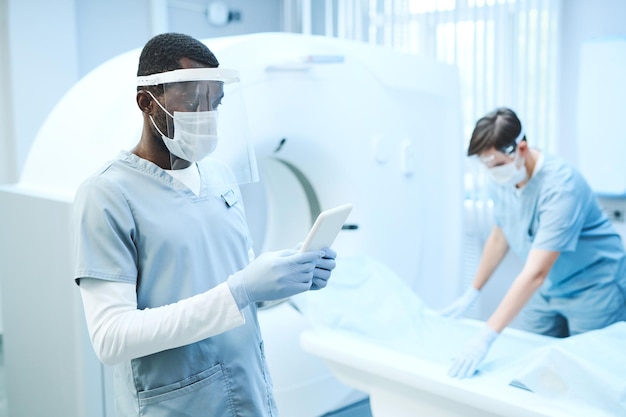 Młody Afroamerykanin w masce i rękawiczkach, korzystający z tabletu w pokoju rezonansu magnetycznego, podczas gdy jego kolega kładzie prześcieradło na stole