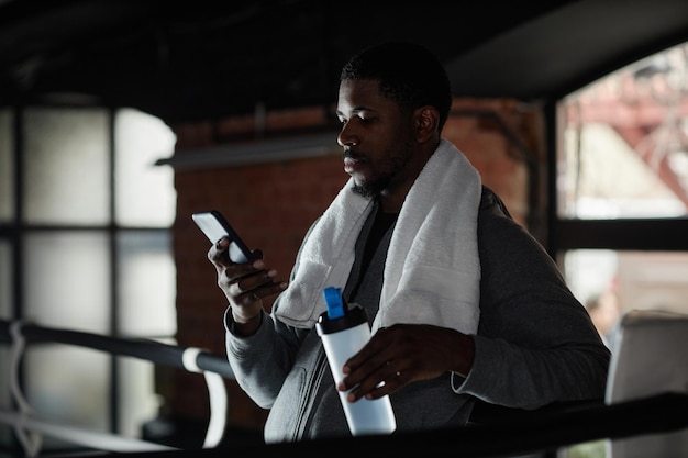 Młody afroamerykański sportowiec z białym ręcznikiem na szyi używający smartfonu