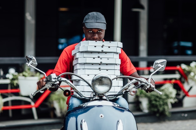 Młody afrykański kurier pozuje na szybkim motocyklu niesie plecak nosi hełm z kartonem