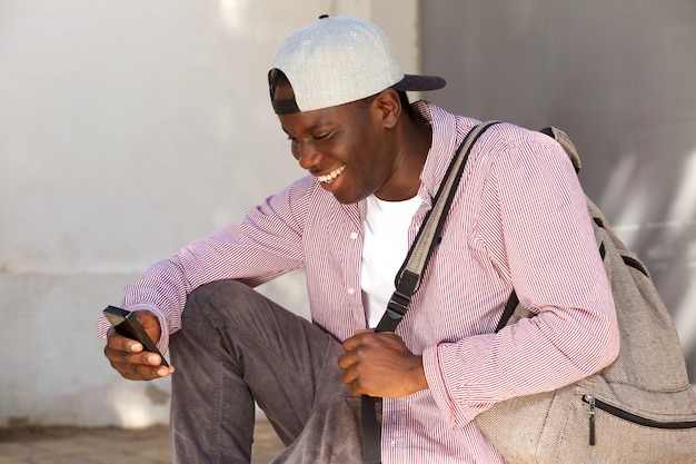 Młody afrykański męski uczeń z nakrętką uśmiechniętą i patrzeje telefon komórkowego