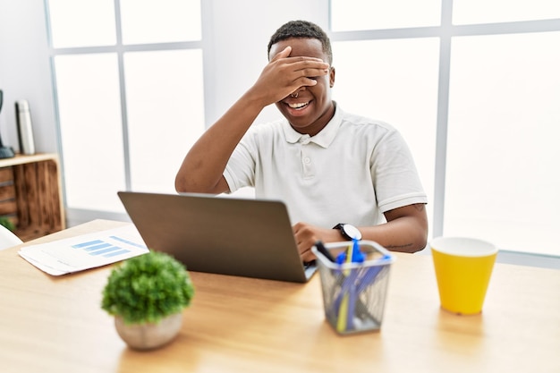Młody afrykański mężczyzna pracujący w biurze za pomocą laptopa komputerowego uśmiechający się i śmiejący się z ręką na twarzy zakrywającą oczy z zaskoczenia. ślepa koncepcja.
