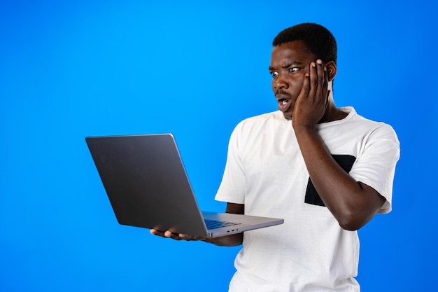 Młody afrykański mężczyzna stoi i używa laptopa na niebieskim tle
