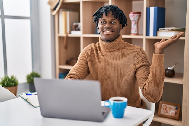 Młody afrykański mężczyzna z dredami pracujący przy użyciu laptopa komputerowego uśmiechający się wesoło, przedstawiający i wskazujący dłonią patrząc w kamerę.