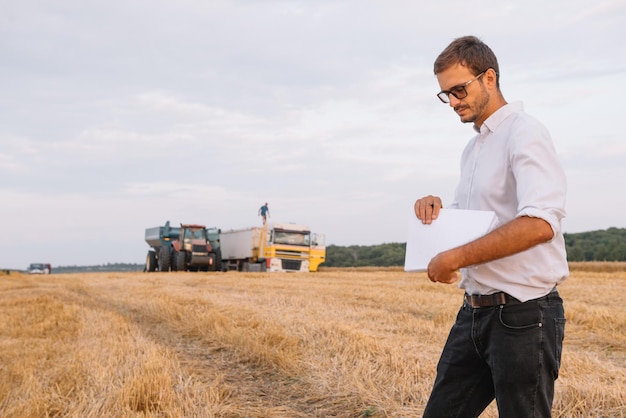 Młody agronom człowiek stojący na polu pszenicy sprawdzanie jakości podczas pracy kombajnu zbożowego.