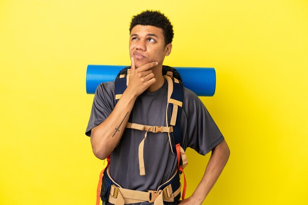 Młody alpinista Afroamerykanin z dużym plecakiem na żółtym tle mający wątpliwości