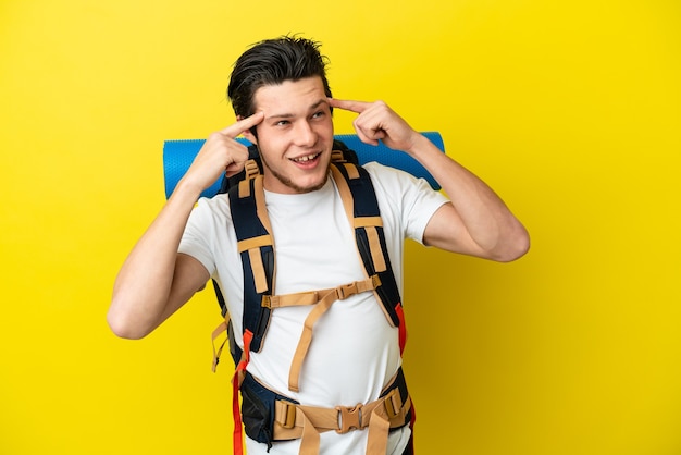 Młody alpinista Rosjanin z dużym plecakiem na żółtym tle mający wątpliwości i myślący