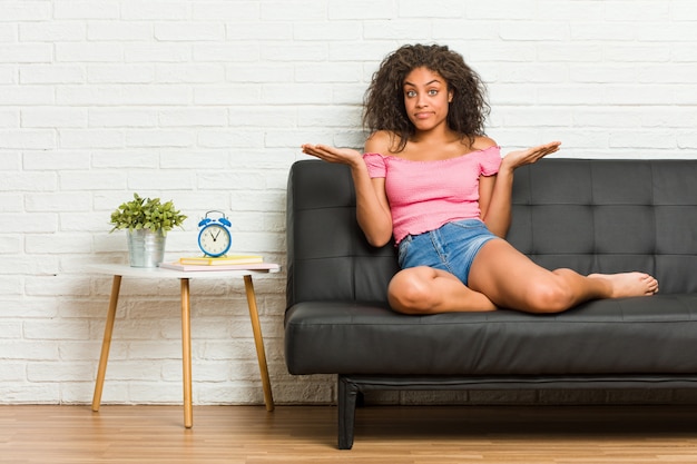 Młody amerykanin afrykańskiego pochodzenia kobiety obsiadanie na kanapie wątpi i wzrusza ramionami ramiona w przesłuchanie gescie.