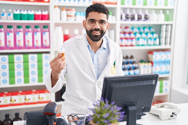 Młody arab farmaceuta za pomocą komputera trzymającego butelkę pigułek w aptece