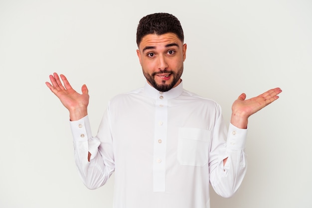 Młody Arab ubrany w typowe arabskie ubrania na białym tle zdezorientowany i wątpliwy wzruszając ramionami, aby trzymać kopię przestrzeni.