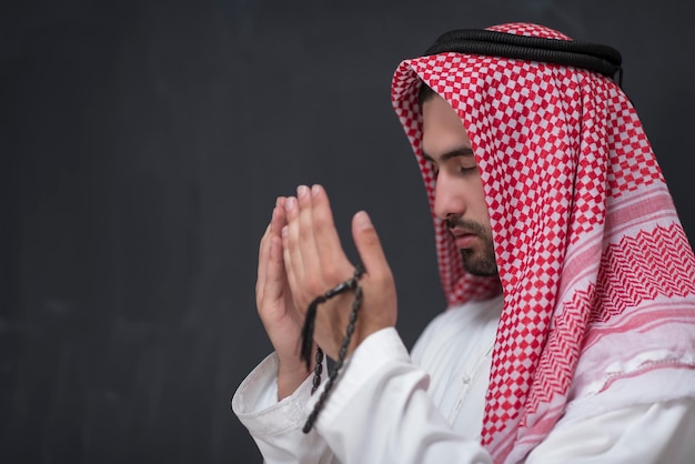 Młody Arab w tradycyjnym stroju, który odmawia tradycyjną modlitwę do Boga, trzyma ręce w geście modlitwy przed czarnym tłem. Wysokiej jakości zdjęcie