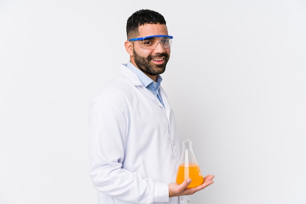 Młody arabski chemik na białym tle wygląda na bok uśmiechnięty, wesoły i przyjemny.
