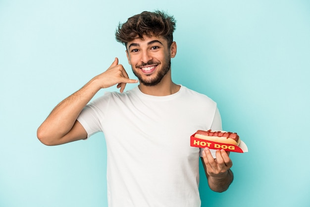 Młody arabski mężczyzna trzyma hotdoga na białym tle na niebieskim tle pokazując gest połączenia z telefonu komórkowego palcami.