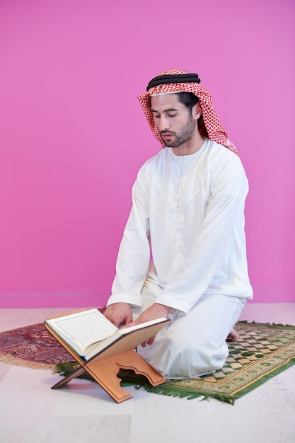 Młody arabski muzułmanin w tradycyjnym stroju czyta świętą księgę Koran na modlącym się dywanie przed różową ścianą przed obiadem iftar podczas uczty ramadanu w domu