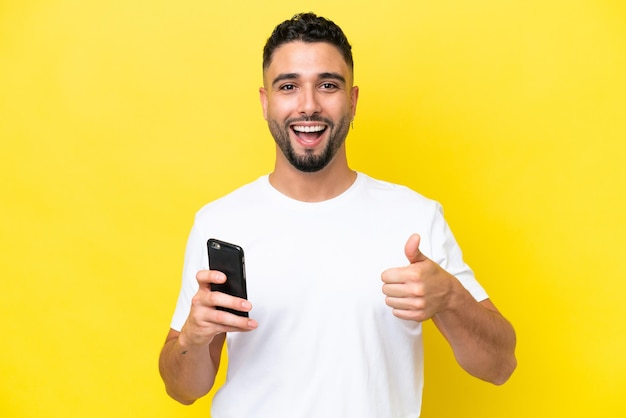 Młody arabski przystojny mężczyzna odizolowany na żółtym tle za pomocą telefonu komórkowego podczas robienia kciuków