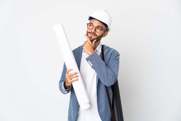 Młody architekt mężczyzna z kaskiem i trzymając plany na białym tle na białej ścianie patrząc uśmiechnięty