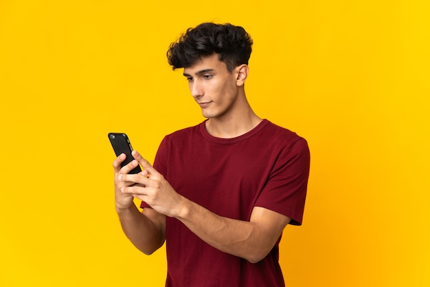 Młody Argentyńczyk na białym tle na żółtej ścianie, wysyłając wiadomość lub e-mail z telefonu komórkowego